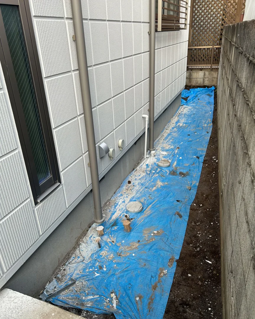 建物側面（犬走り）の施工前の様子。配管の周りにブルーシートが敷かれ、これから整備が行われる土のエリア。