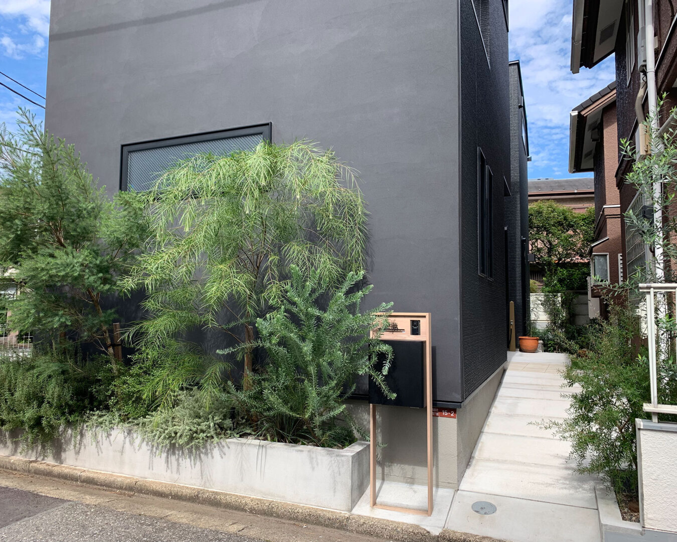 新築の黒い住宅の前に施工された、コンクリート花壇の豊かな植栽とオリジナルポスト、奥へ続くモルタルスロープ。