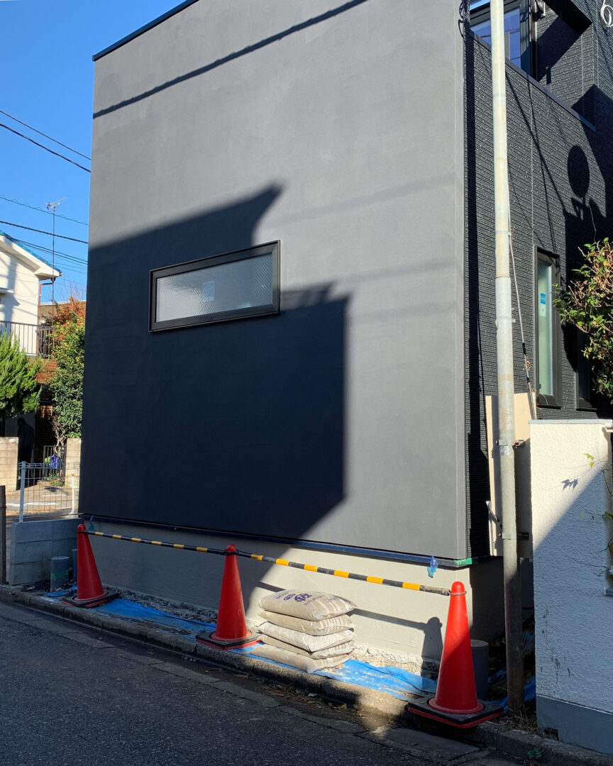 外構着工前の様子。黒い新築住宅の前面道路沿いにカラーコーンが並び、基礎部分の工事が進められている状態。