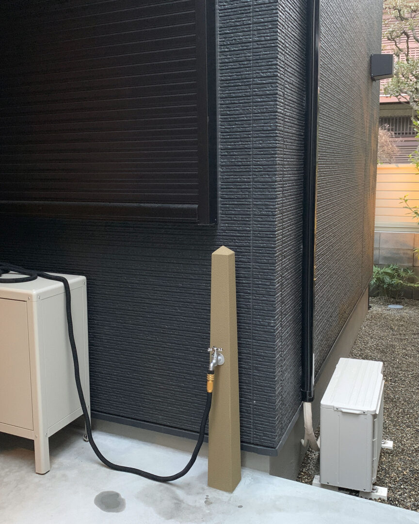 黒い外壁のコーナー部分にスッキリと設置された、スタイリッシュなブラウンのデザイン立水栓と屋外設備。